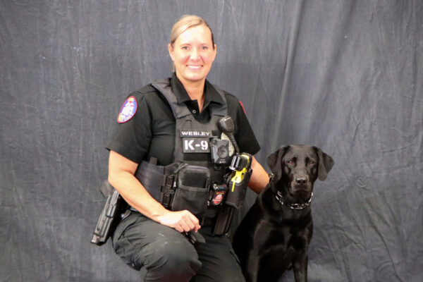 Police Officer Wesley, wearing a black police uniform, poses with canine Uman, a black labrador retriever